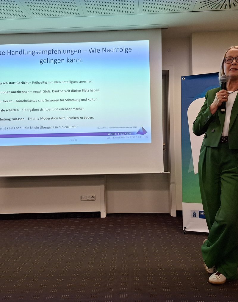 Unternehmensberaterin Anke Tielker steht gelehnt an einem Stehtisch und im Hintergrund ist die Projektion einer ihrer Vortragsfolien zu sehen.