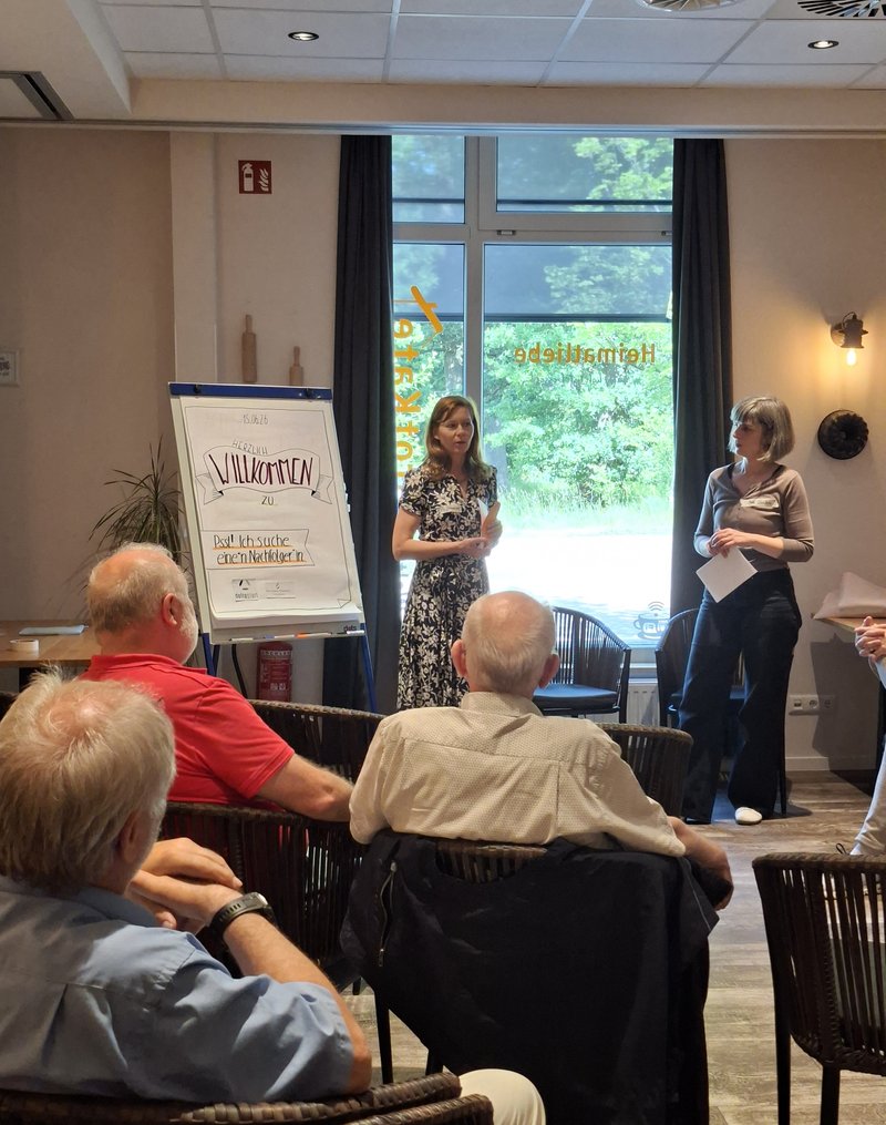 2 Personen neben einem Flipchart in einer Vortragssituation, einige Köpfe aus dem Publikum von hinten sichtbar