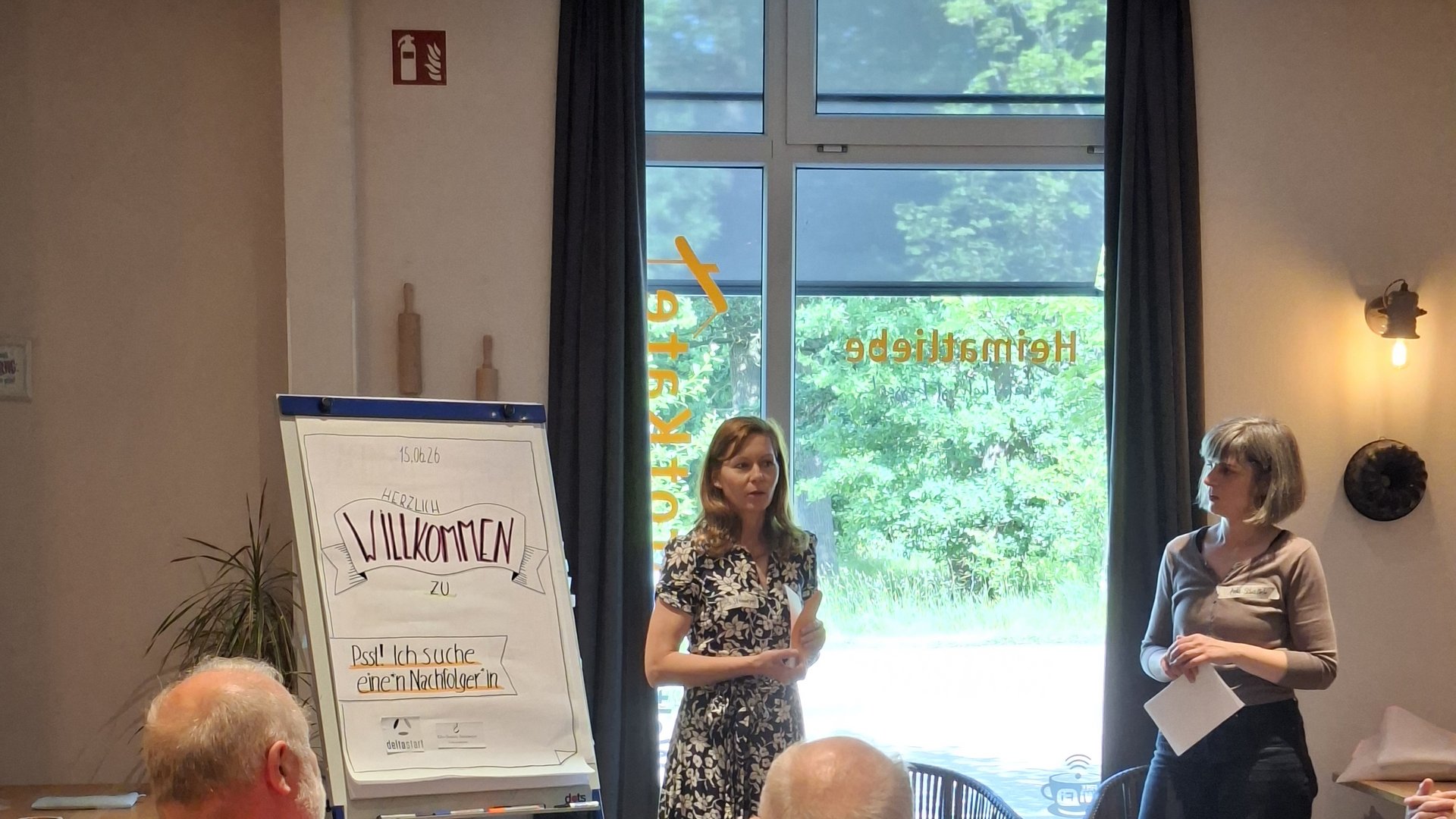 2 Personen neben einem Flipchart in einer Vortragssituation, einige Köpfe aus dem Publikum von hinten sichtbar