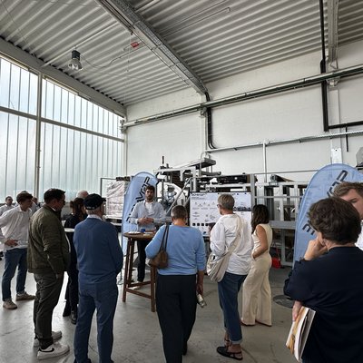 Mehrere Menschen von hinten, die in Richtung eines eher jüngeren Mannes vor einer Produktionsmaschine, neben dem Mann eine Infotafel. Er ist von vorn zu sehen, teils verdeckt durch sein Publikum, und spricht.
