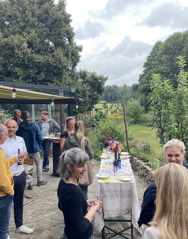 Das Foto zeigt einen Ausschnitt einer Terrasse und einem weitläufigen Garten im Hintergrund. Auf der Terrasse sind mehrere Grüppchen an Menschen, die miteinander im Gesporäch sind.
