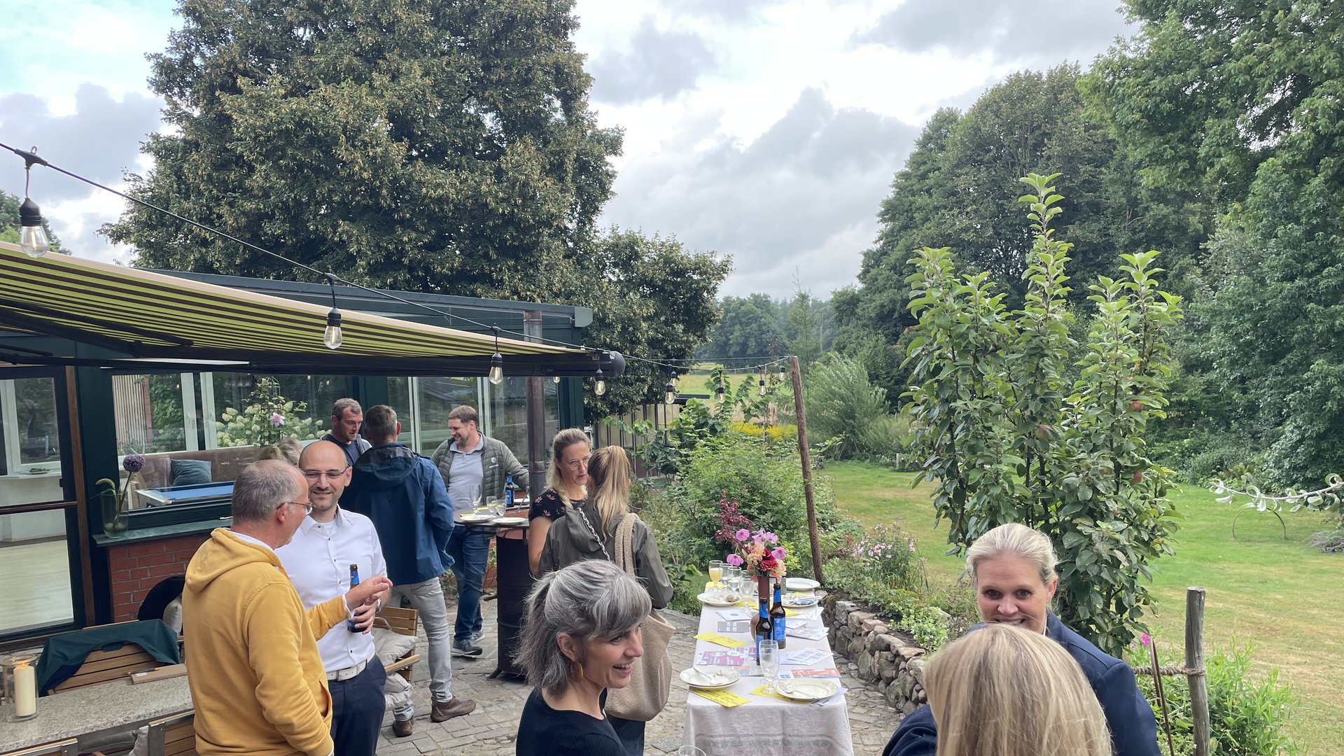 Das Foto zeigt einen Ausschnitt einer Terrasse und einem weitläufigen Garten im Hintergrund. Auf der Terrasse sind mehrere Grüppchen an Menschen, die miteinander im Gesporäch sind.