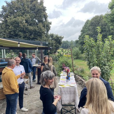 Das Foto zeigt einen Ausschnitt einer Terrasse und einem weitläufigen Garten im Hintergrund. Auf der Terrasse sind mehrere Grüppchen an Menschen, die miteinander im Gesporäch sind.